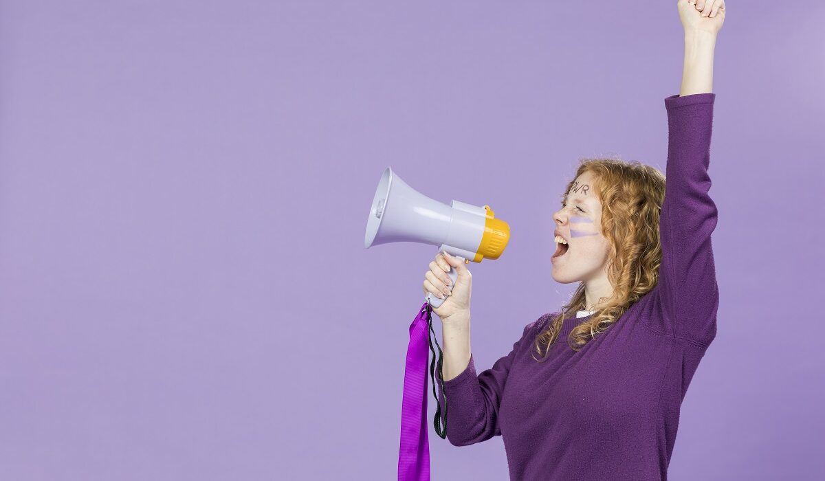 Mlada žena s crvenom kosom stoji ispred ljubičaste podloge i s megafonom u ruci uzvikuje dobre parole i vijesti