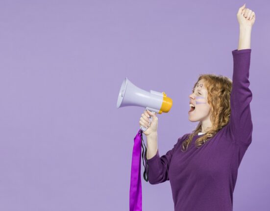 Mlada žena s crvenom kosom stoji ispred ljubičaste podloge i s megafonom u ruci uzvikuje dobre parole i vijesti